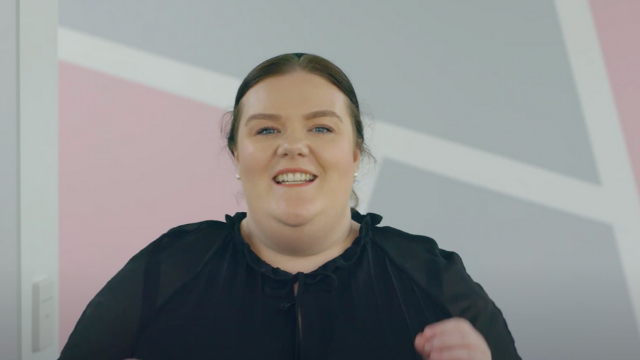 A woman in a black shirt is smiling in front of a pink and gray wall.