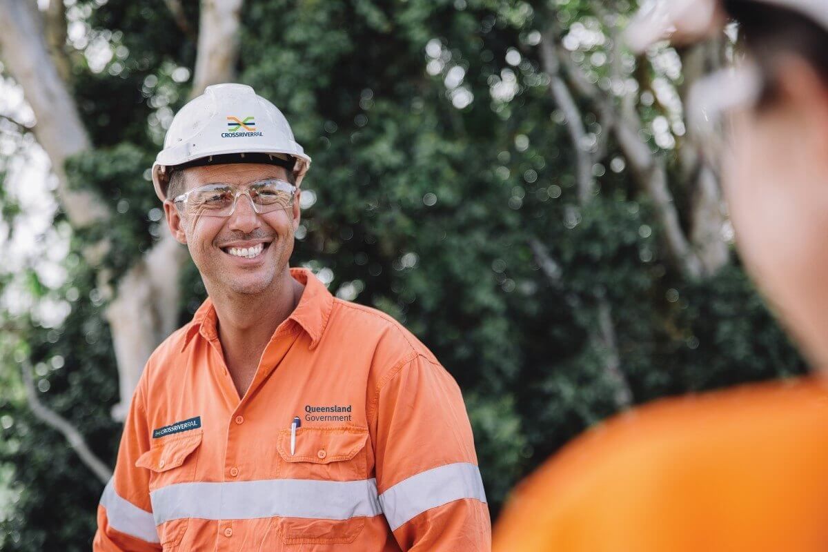 A man wearing a hard hat and safety glasses is smiling while standing next to another man.