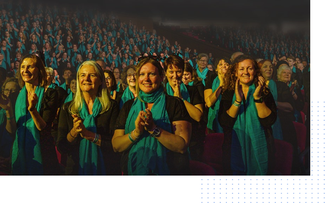A group of women wearing teal scarves are standing in front of a crowd of people applauding.