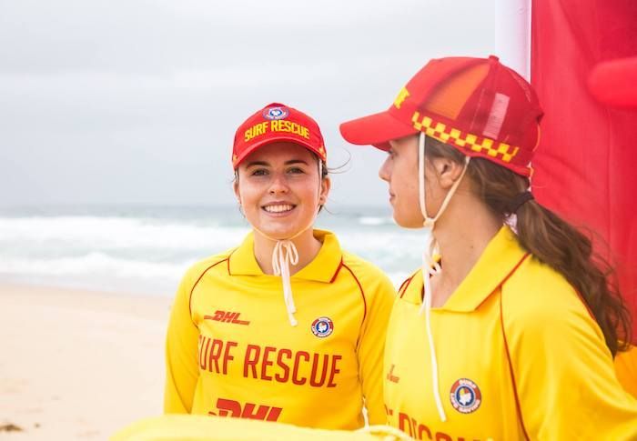 Two lifeguards wearing yellow shirts and red hats are standing on a beach.