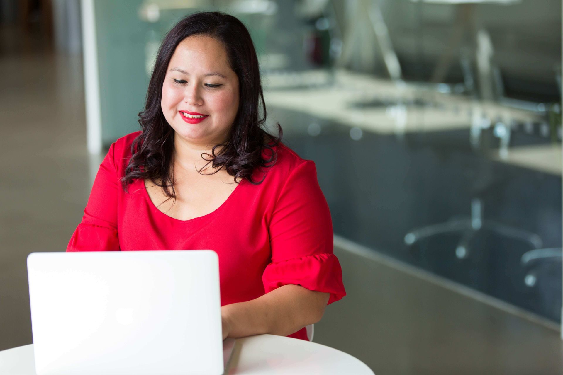 A woman in a red shirt is sitting at a table using a laptop computer.