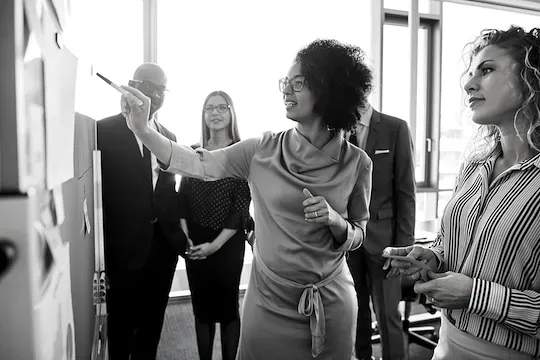 A black and white photo of a group of people standing around a whiteboard.