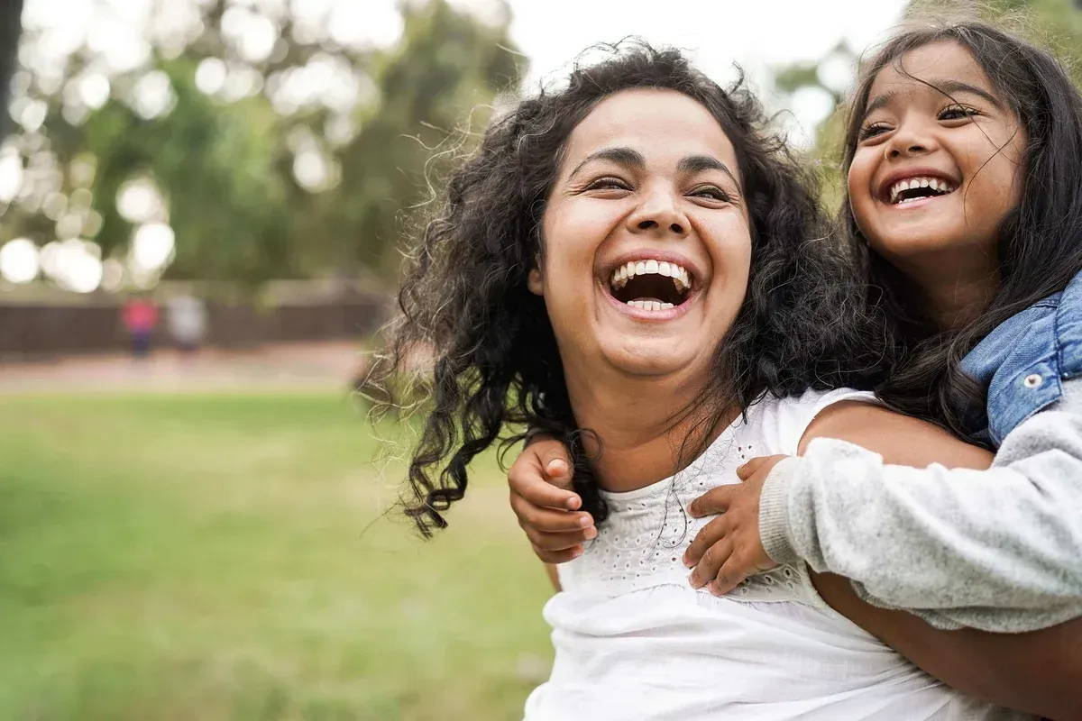 A woman is carrying a little girl on her back in a park.