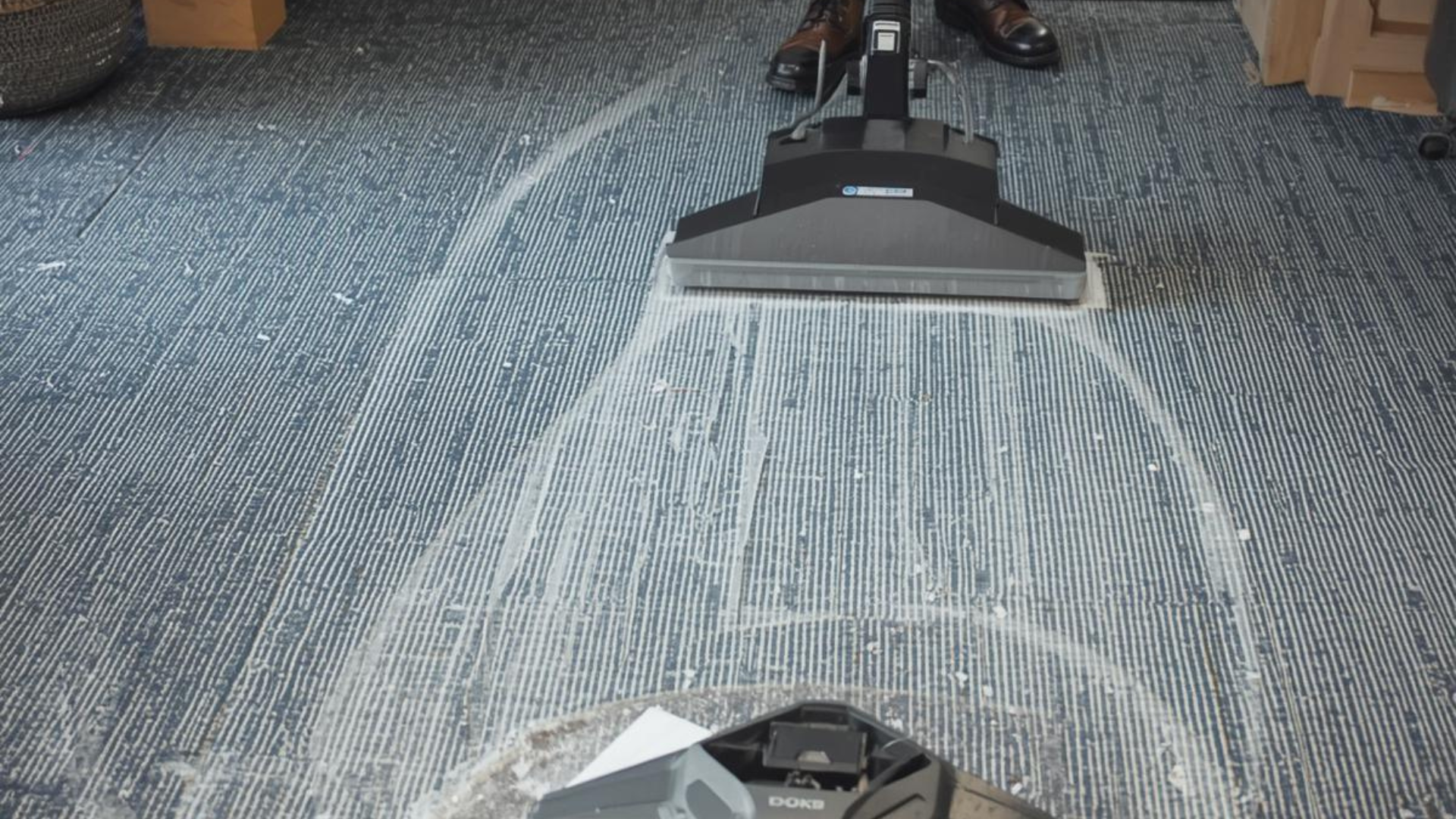 Vacuum cleaner cleaning a blue carpet, creating lighter streaks. A person's legs and feet are visible.