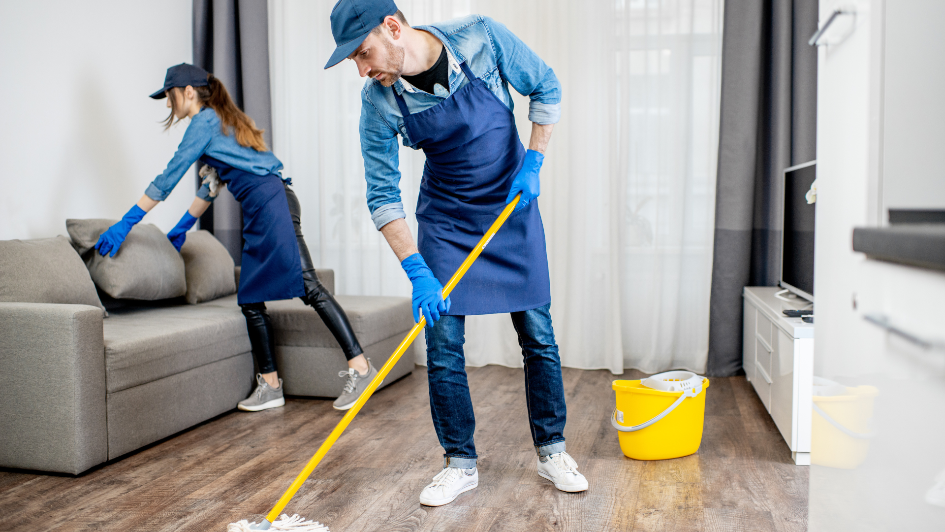 Two people in uniform cleaning a living room: one mopping, the other arranging a couch cushion.