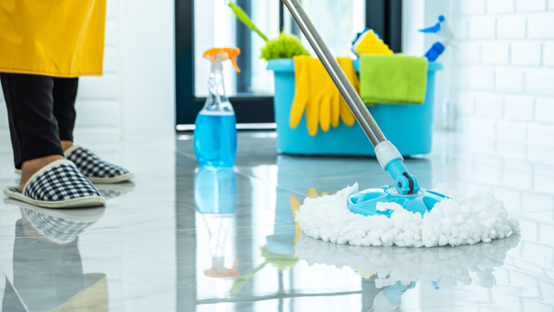 Person mopping a shiny, white tiled floor with cleaning supplies in a blue bucket nearby.