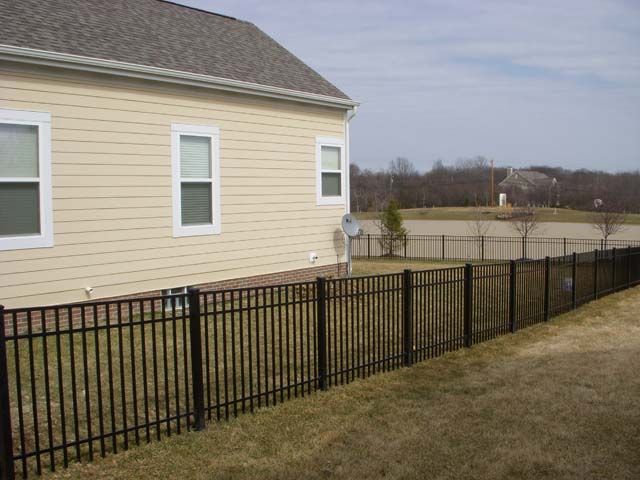 A new wooden privacy fence stands in a grassy yard in front of a two-story gray house under a blue sky.