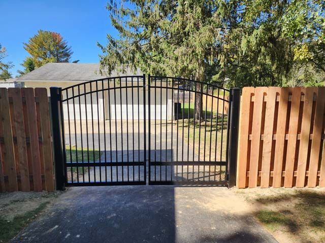 A black metal driveway gate stands between two brick pillars with mounted lanterns in front of a house.