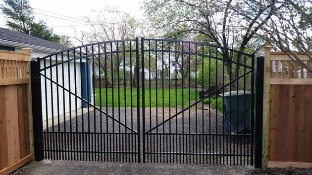 Black decorative iron double gate set between two beige brick pillars topped with lanterns, framing a house and garden.