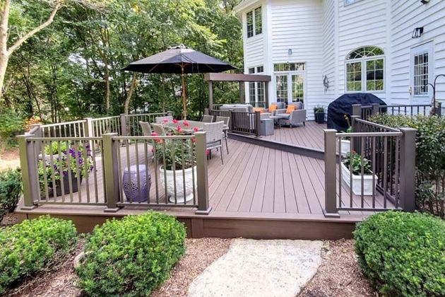 A wooden pergola with a lattice roof sits on a square deck in a backyard next to a brick house.