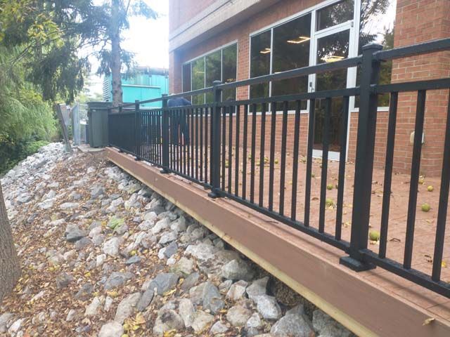 Close-up of a brown wooden deck railing with a vertical post and diagonal support, set against a blurred backyard.