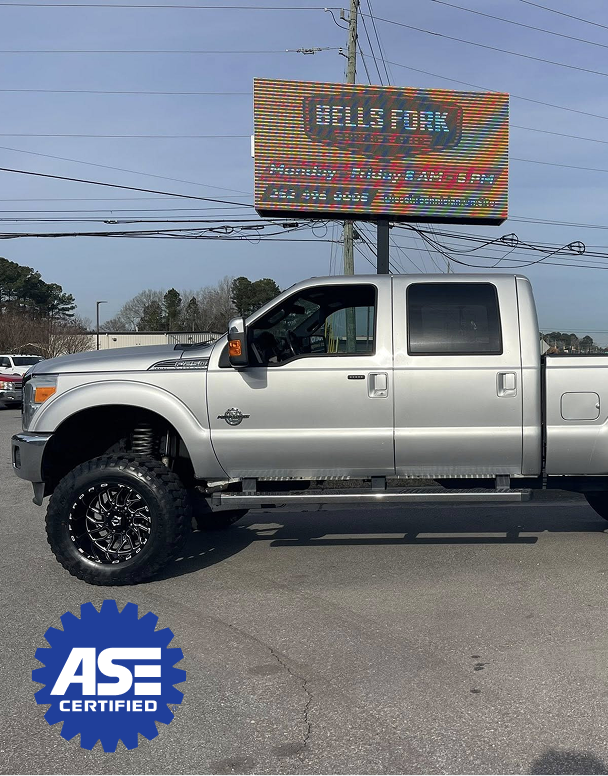 Silver lifted pickup truck in front of Bells Ford auto shop. ASE certified sticker visible.