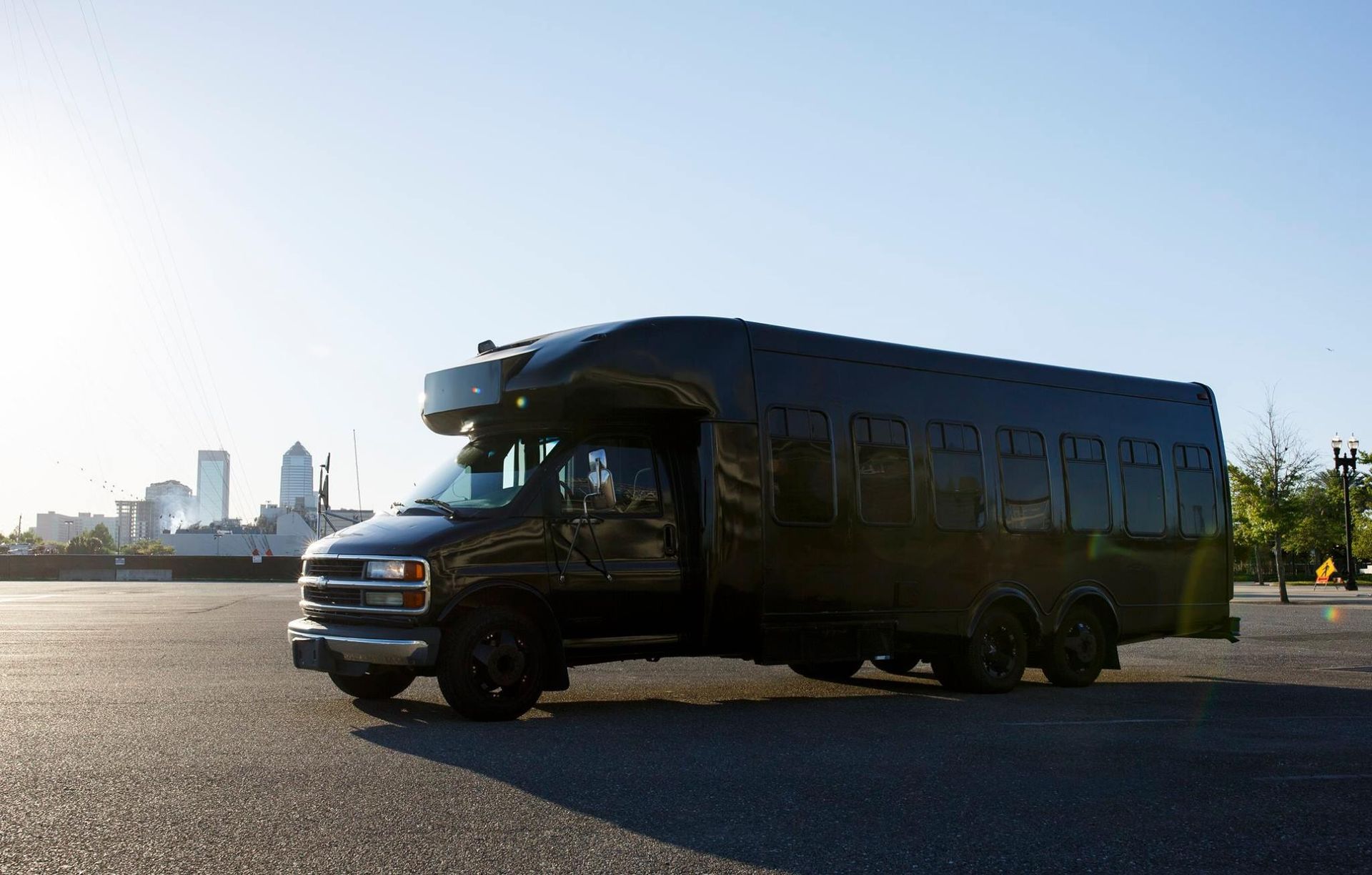 Black bus/van parked on a paved lot with city skyline in the background at sunset
