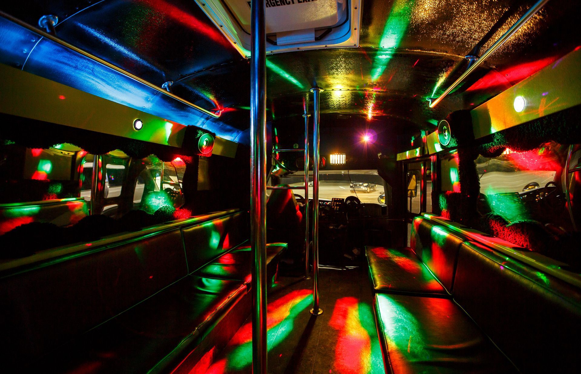 Inside a dim party bus with colorful neon lights reflecting on metal poles and benches