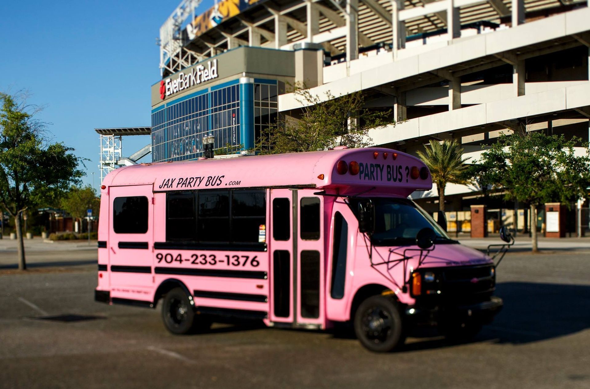 Pink shuttle bus parked outside a stadium, with black lettering and phone number on the side.