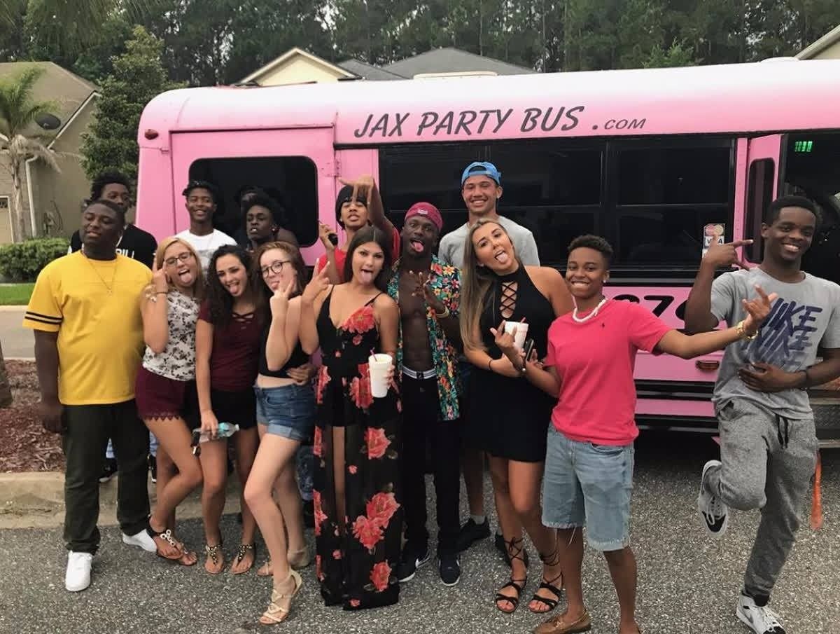 Group posing in front of a pink “JAX PARTY BUS” with smiling, colorful outfits outdoors