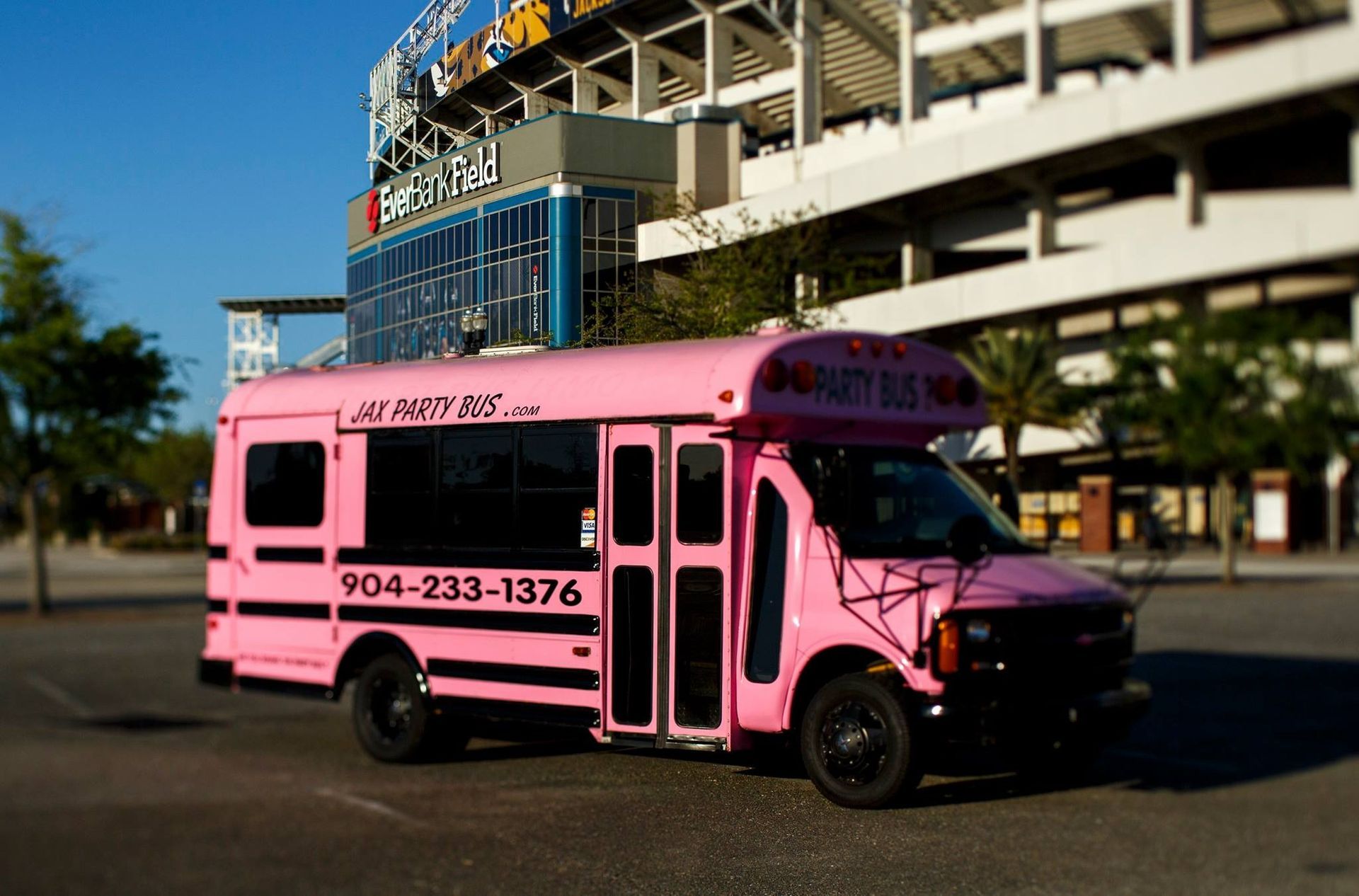 Pink shuttle bus parked outdoors near a stadium, with a blue glass entrance in the background