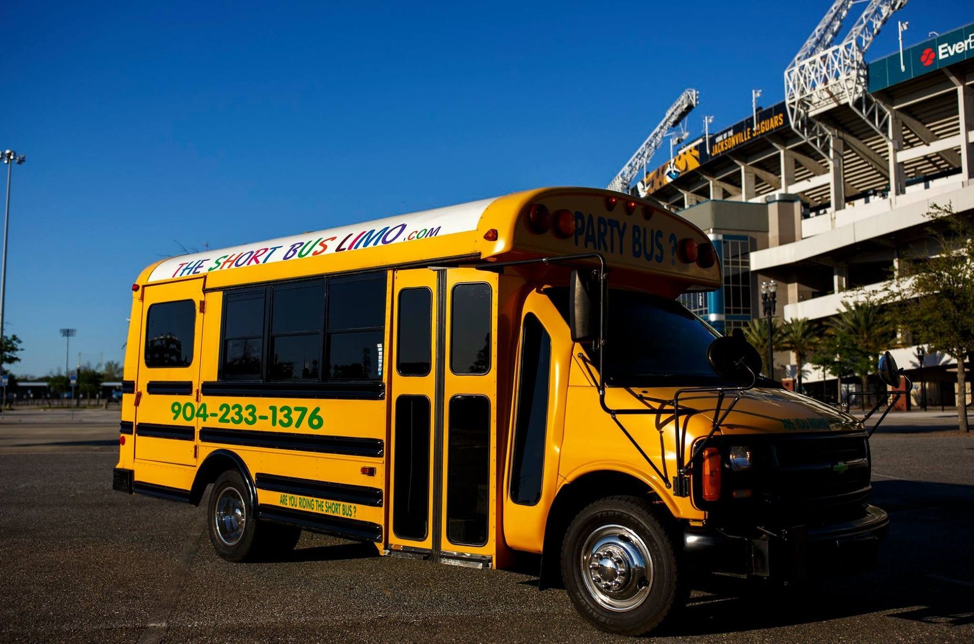 Yellow school bus parked outside a stadium, with blue lettering and a visible phone number.