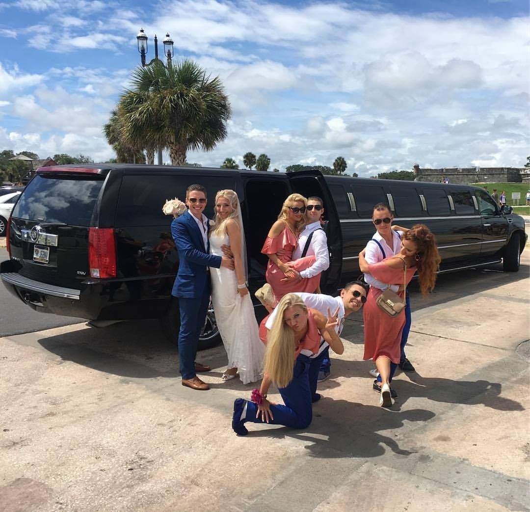 Group posing and dancing beside black limousines on a sunny street, with one person kneeling in front
