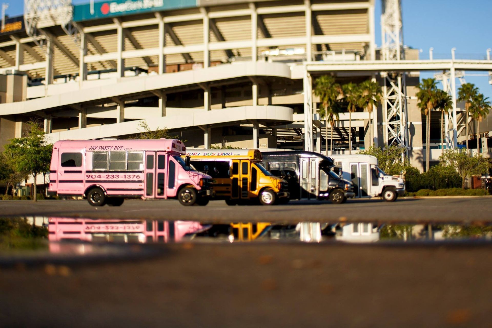 Pink bus and parked trucks in front of a stadium, reflected in a puddle