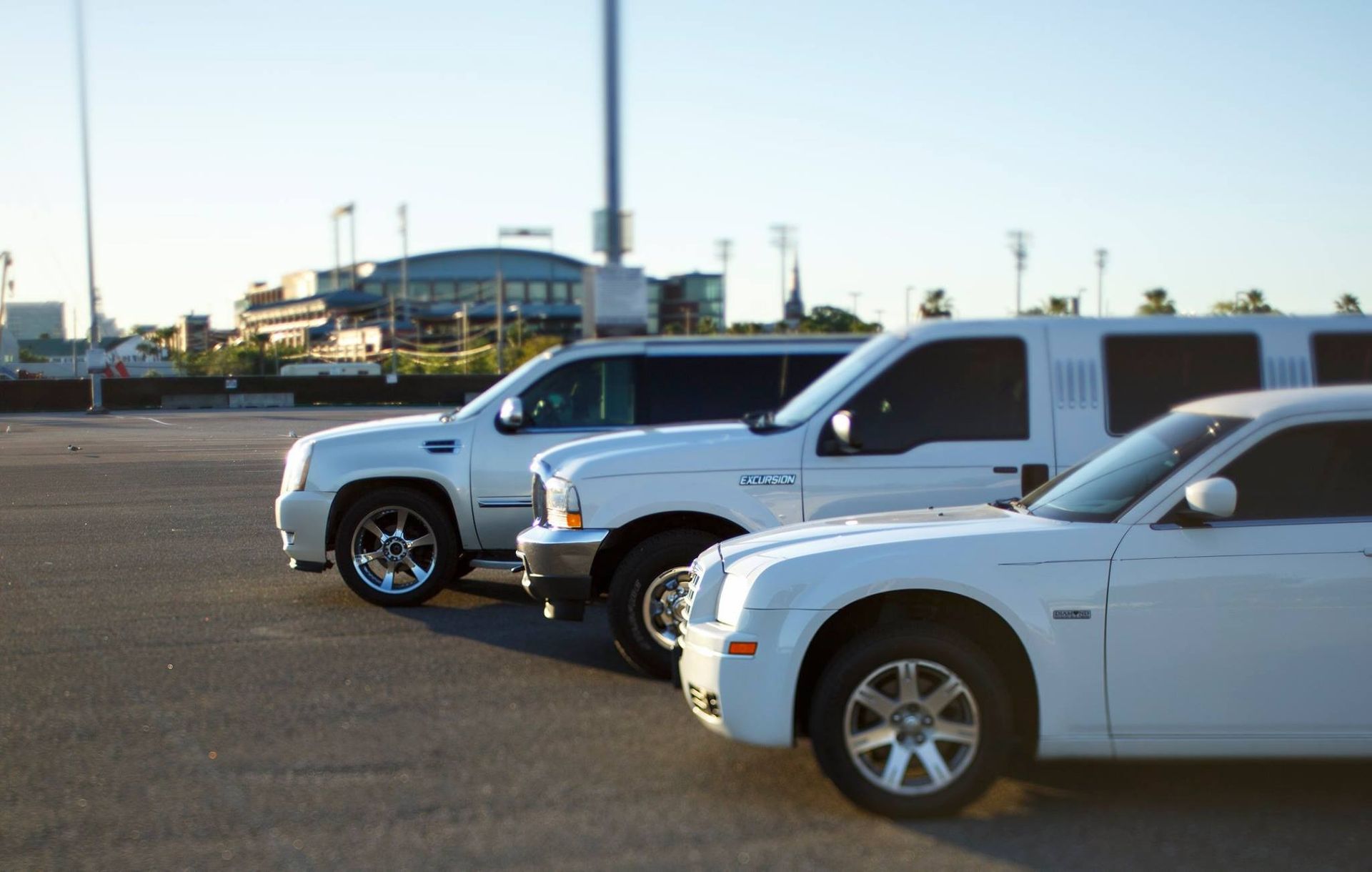Three white pickup trucks parked side by side in a lot with a stadium in the background