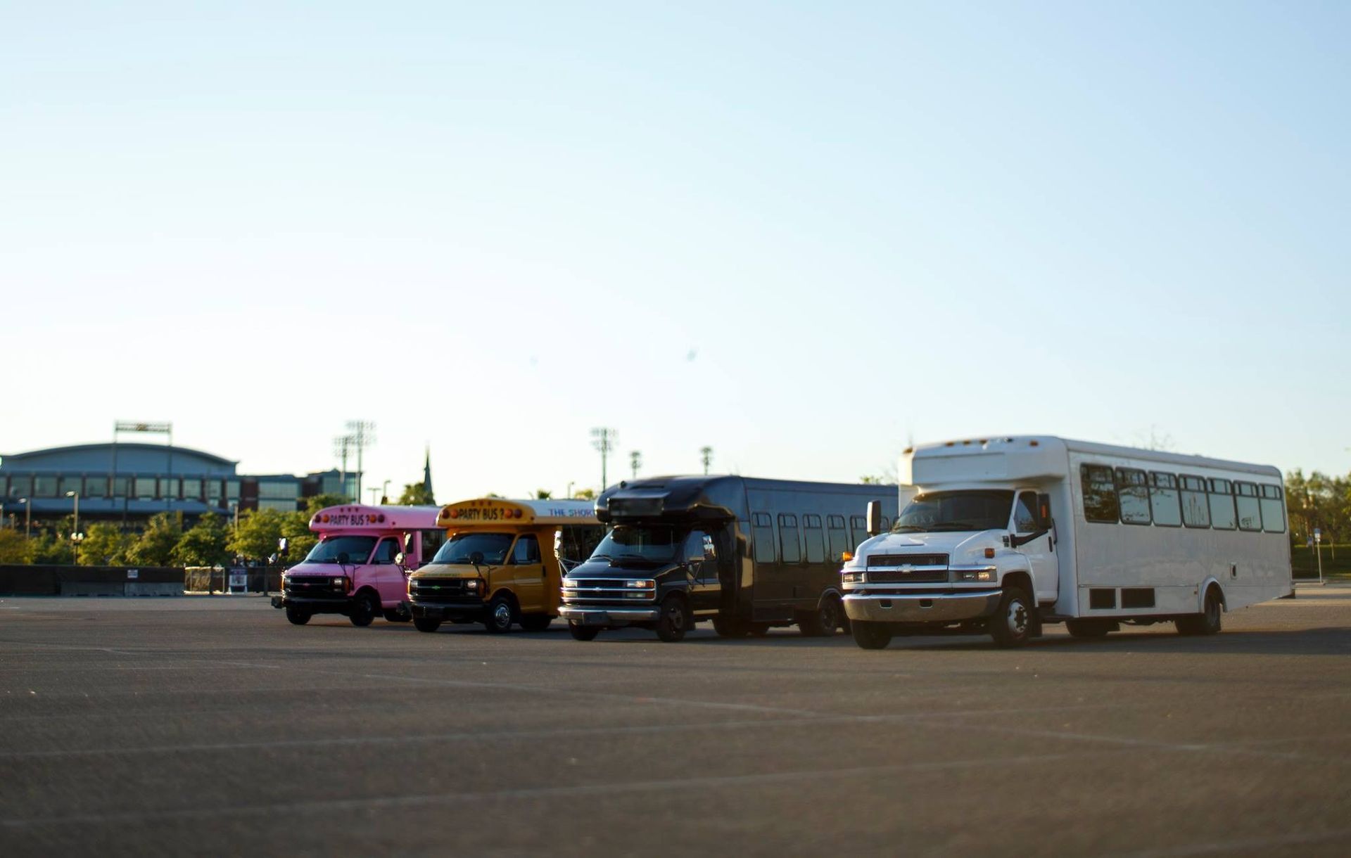Four colorful buses and a white shuttle parked in a row on an open lot under a clear sky