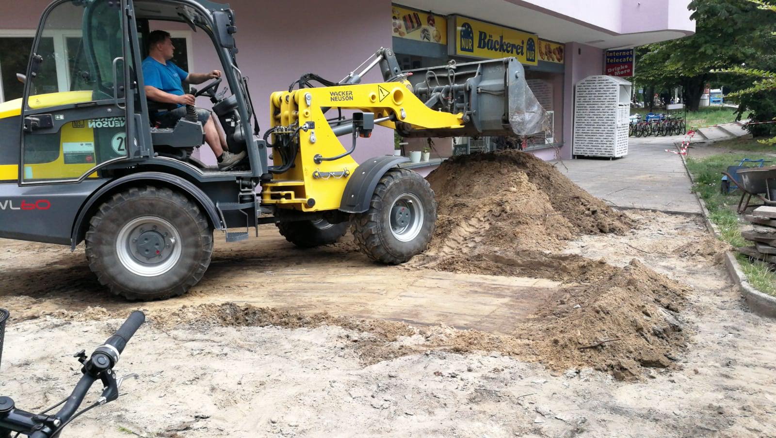 Ein Mann bedient einen gelb-grauen Minilader und bewegt Erde in der Nähe des Eingangs eines Gebäudes.