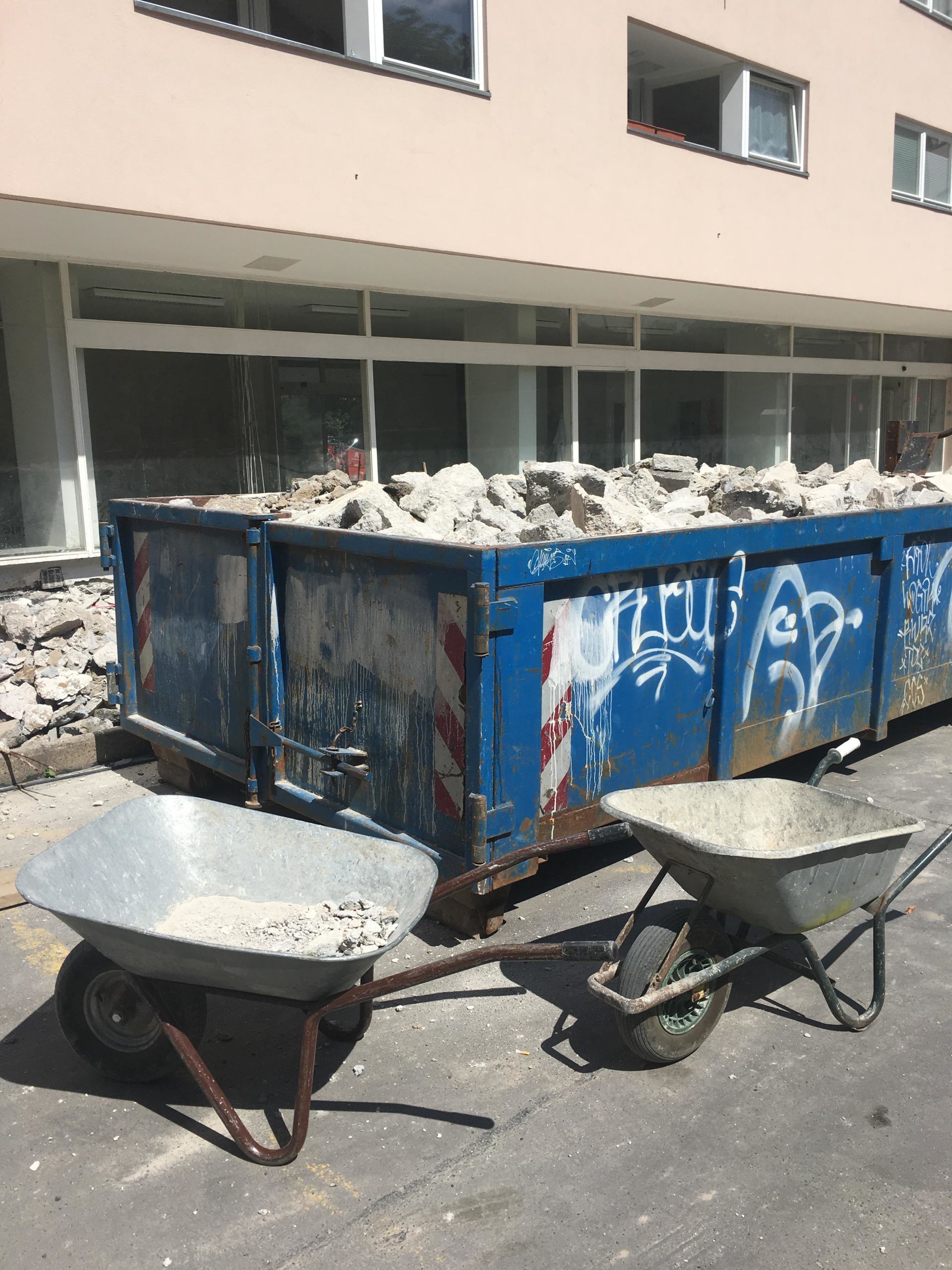 Blauer Müllcontainer voller Schutt, zwei Schubkarren und ein Gebäude im Hintergrund.