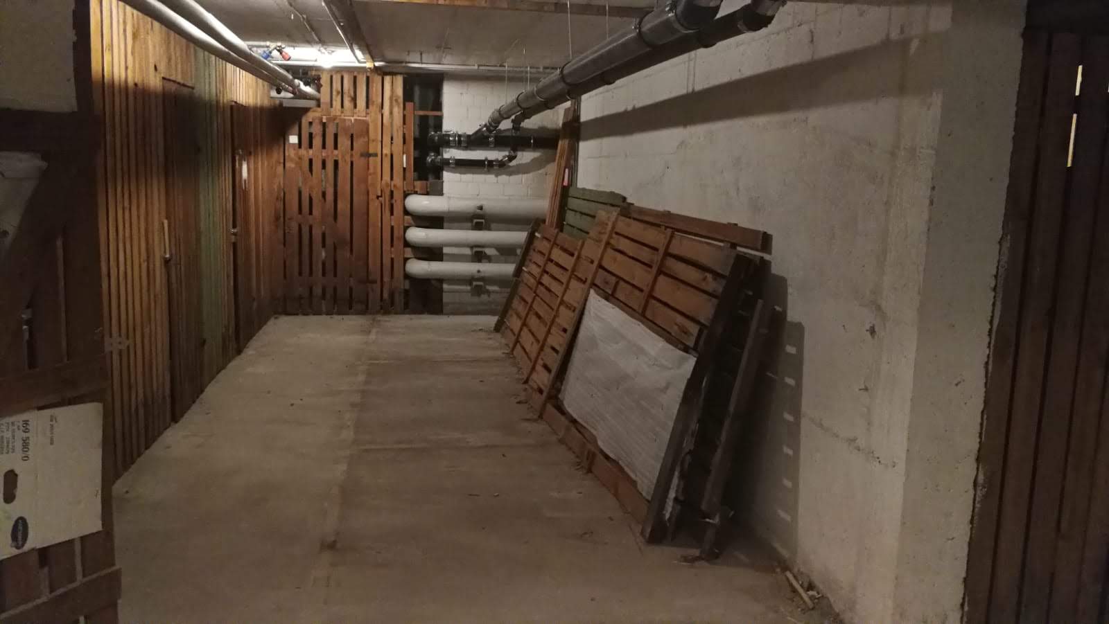 Schmaler Betonflur mit Holzvertäfelung, Rohren und an der Wand lehnenden Paletten.
