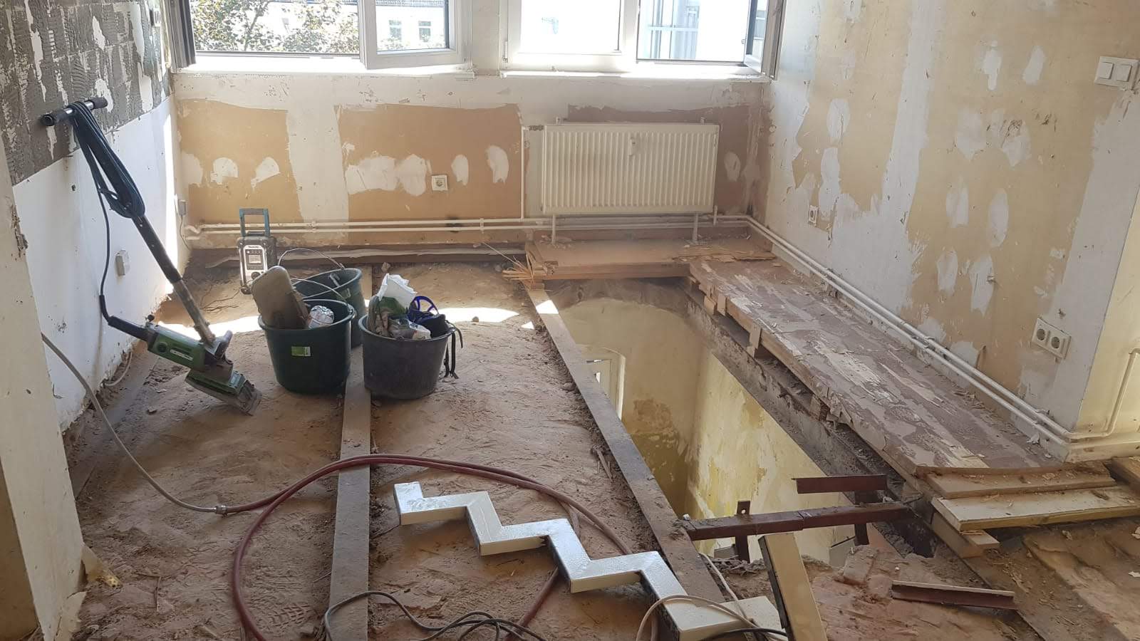 Zimmer wird renoviert: freiliegender Bodenbelag, beschädigte Wände, Bauwerkzeuge und ein Heizkörper.