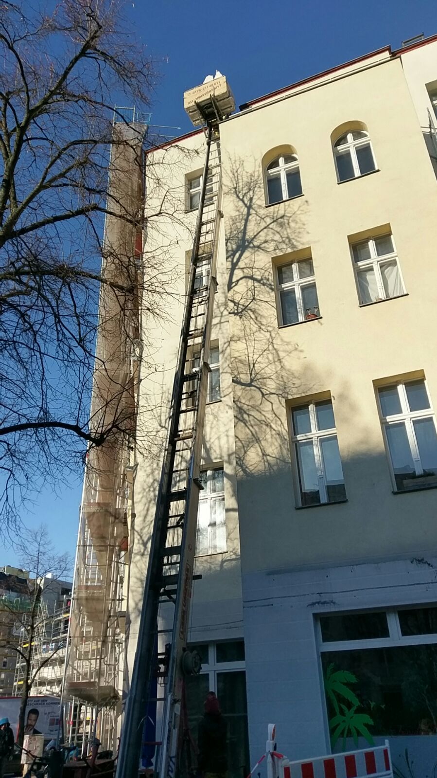 Ein Aufzug hebt eine Kiste in die oberen Stockwerke eines Gebäudes; Szene bei Tageslicht vor einem hellen Wohnhaus.