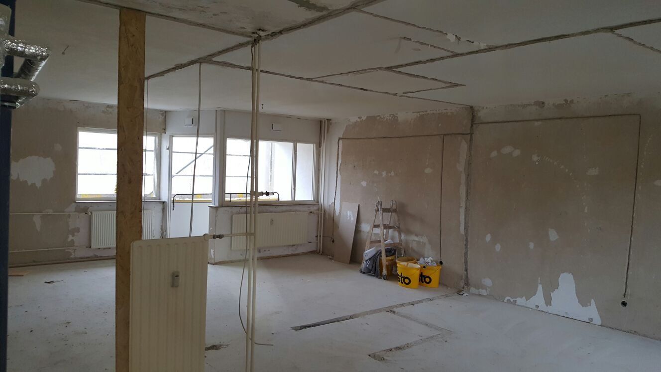 Innenansicht eines im Bau befindlichen Raums mit freiliegenden Kabeln, unfertigen Wänden, Fenstern und Baumaterialien.