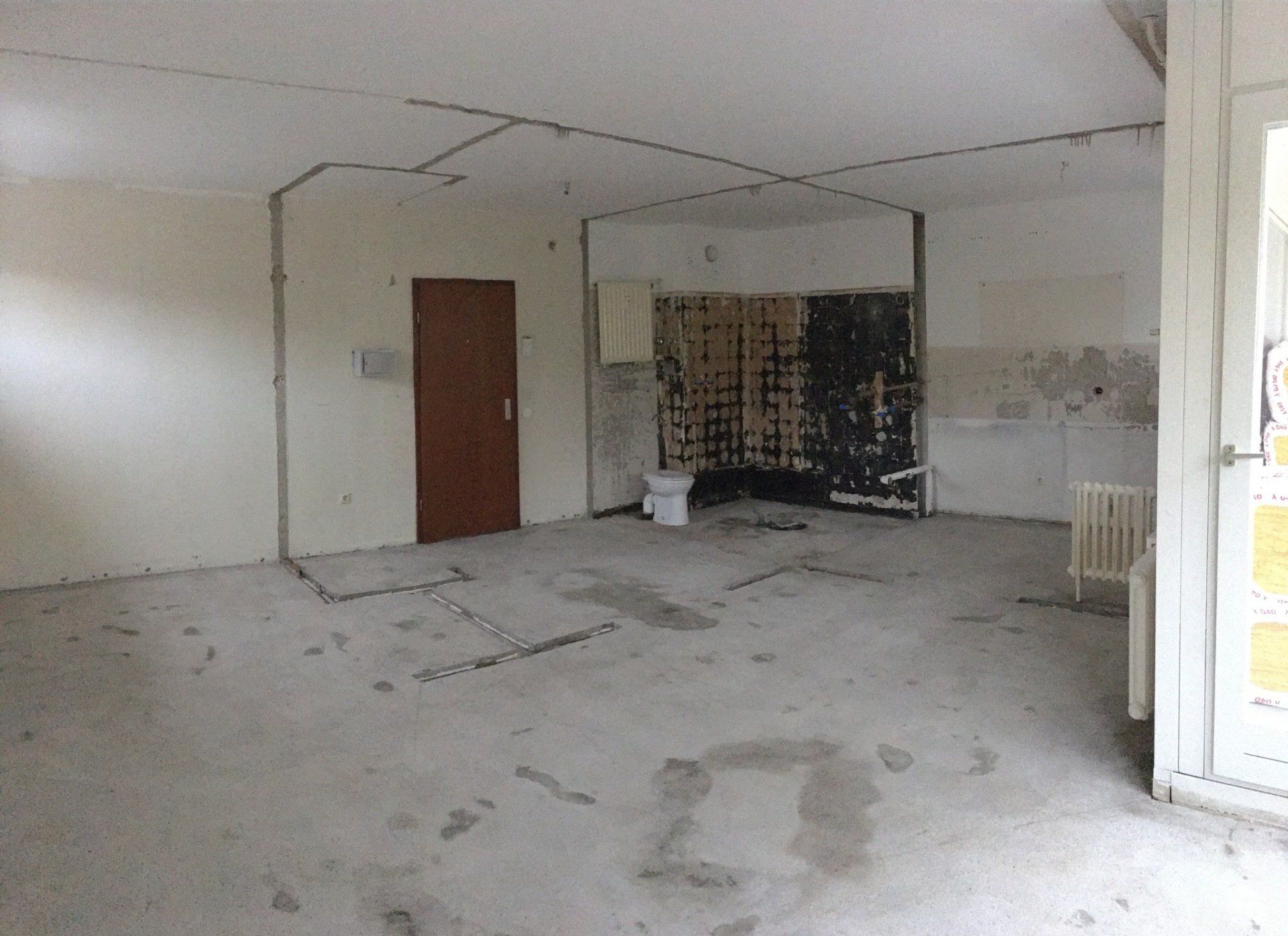 Leerer Raum wird renoviert. Wände und Boden bestehen aus nacktem Beton. An einer Wand befindet sich eine braune Tür.