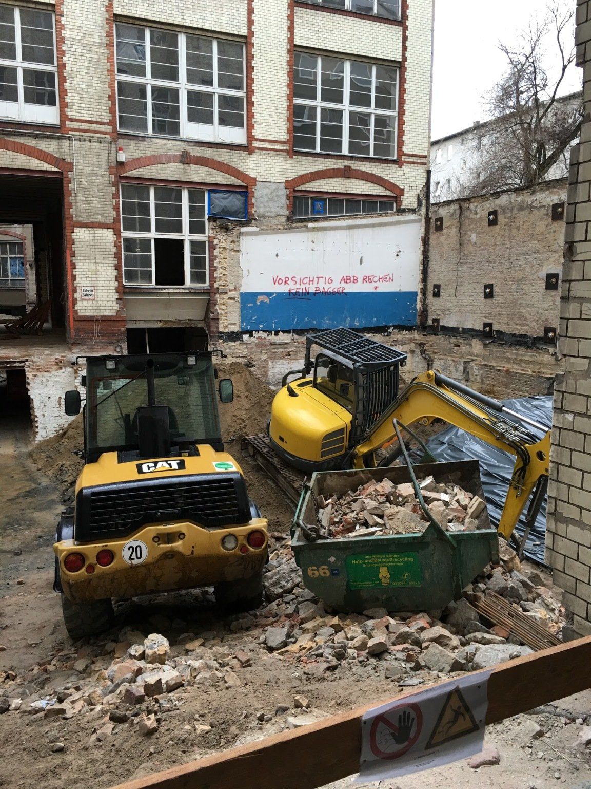 Baustelle mit einem gelben Bagger, der einen grünen Container befüllt, in der Nähe eine gelbe CAT-Maschine.