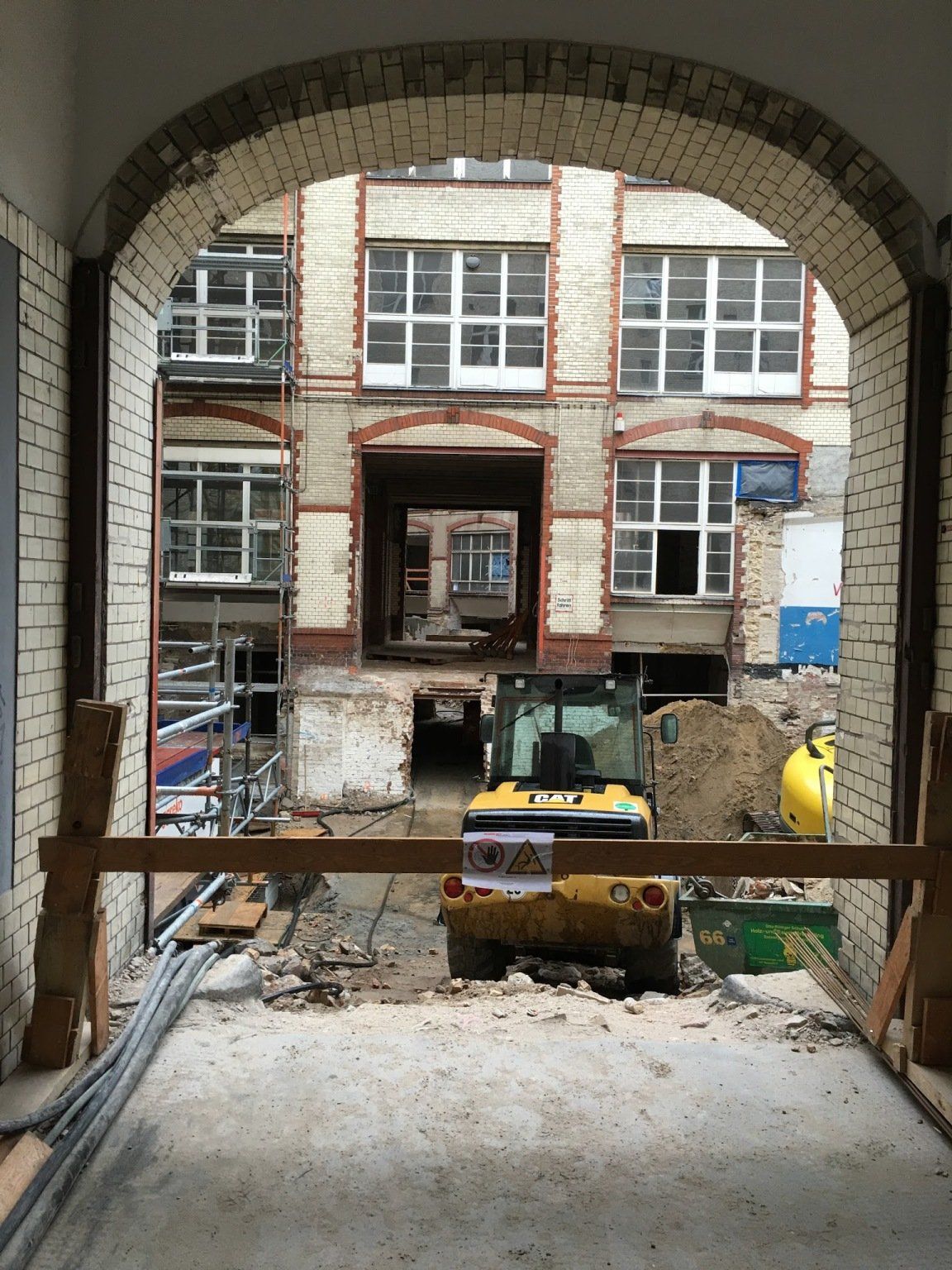 Blick durch einen Torbogen auf eine Baustelle mit einem Minibagger vor einem Backsteingebäude.