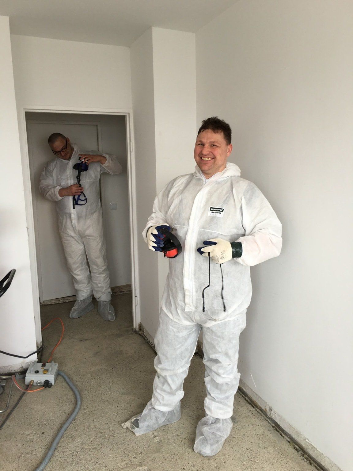 Zwei Männer in weißen Schutzanzügen in einem Raum, einer lächelt und hält Werkzeuge, der andere steht in einem Türrahmen.