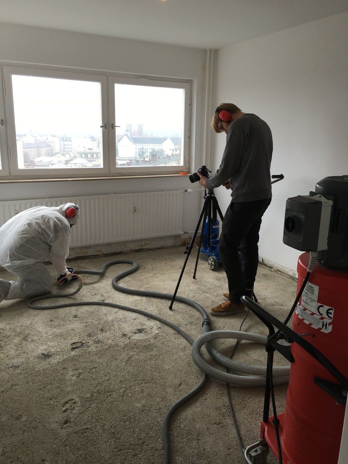 Zwei Arbeiter in einem Raum. Einer filmt mit einem Stativ, der andere arbeitet im Schutzanzug auf dem Boden.