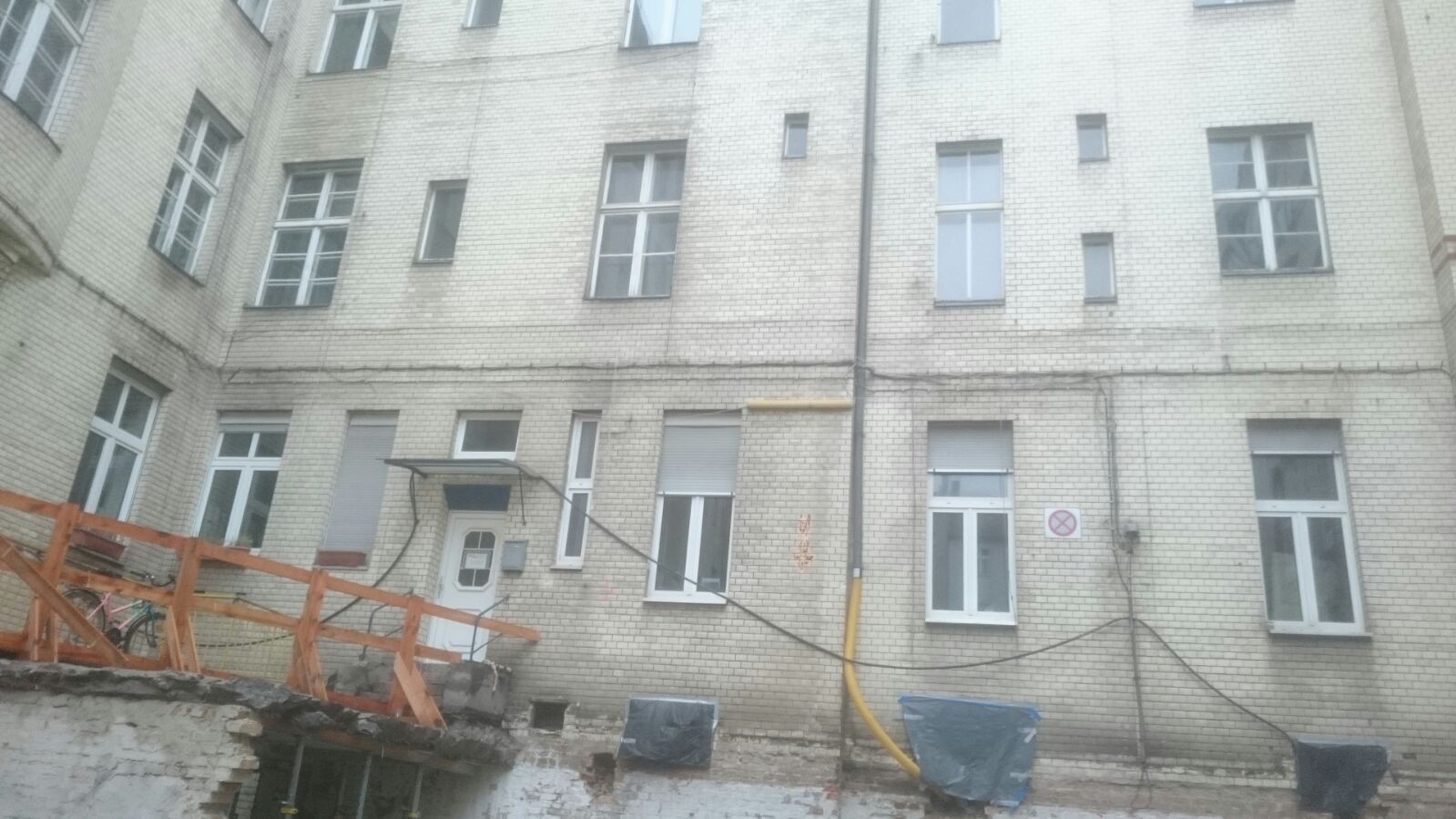 Außenansicht eines Backsteingebäudes mit mehreren Fenstern, einige mit Jalousien. Orangefarbenes Geländer im Vordergrund.