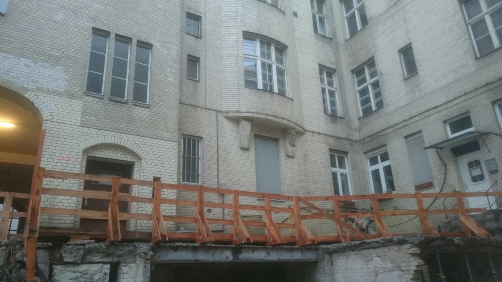 Gebäudehof mit im Bau befindlicher Brücke; cremefarbene Ziegelwände, Holzgeländer, bedeckter Himmel.