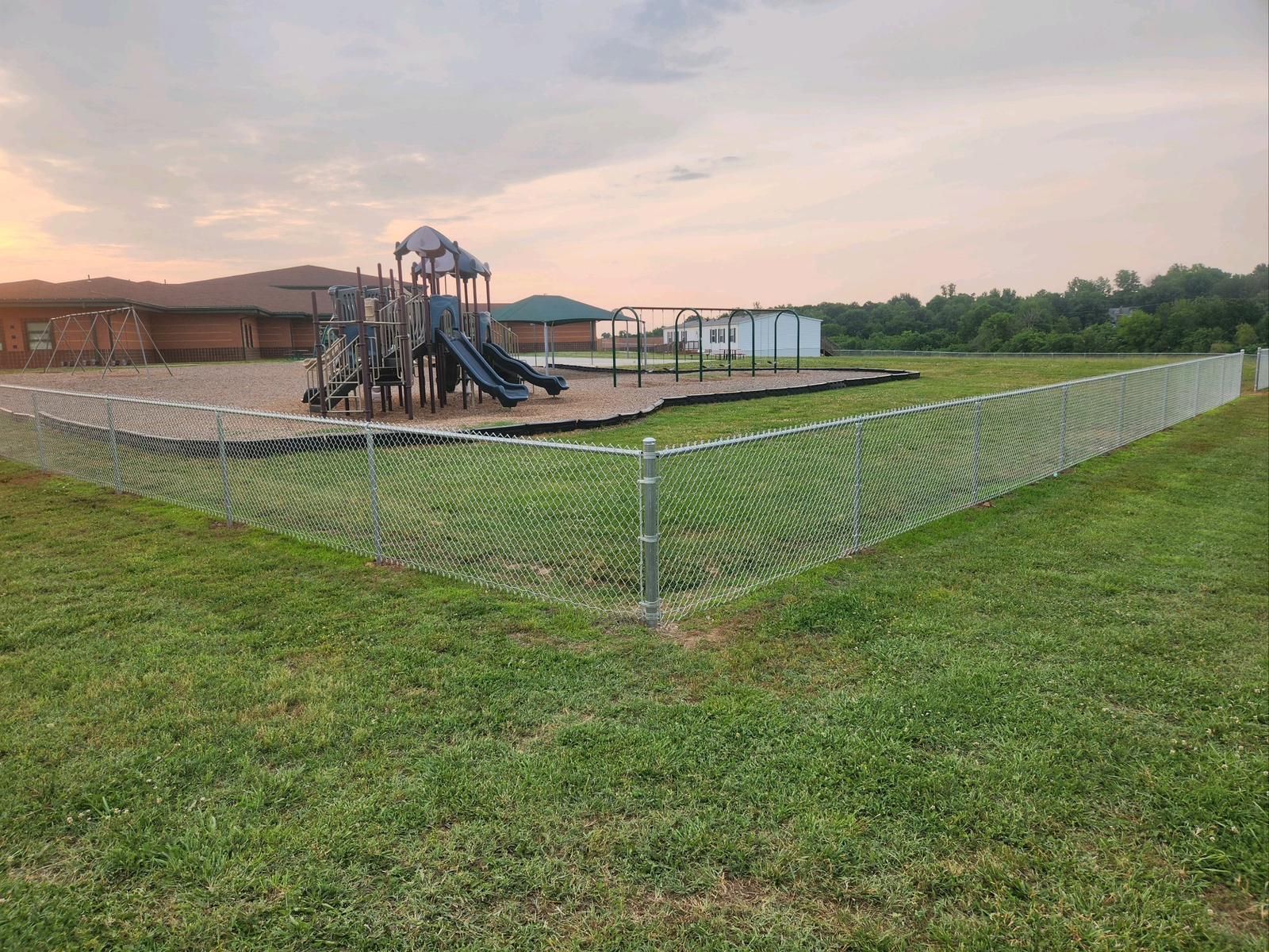 A chain link fence surrounds a playground in a grassy field.