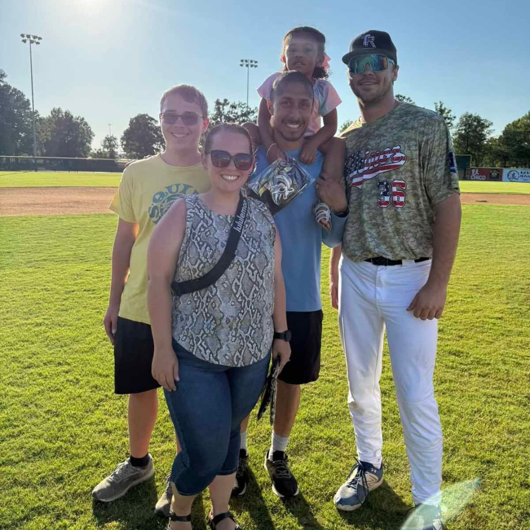 Five people posing together on a sunny baseball field, smiling near the outfield grass.
