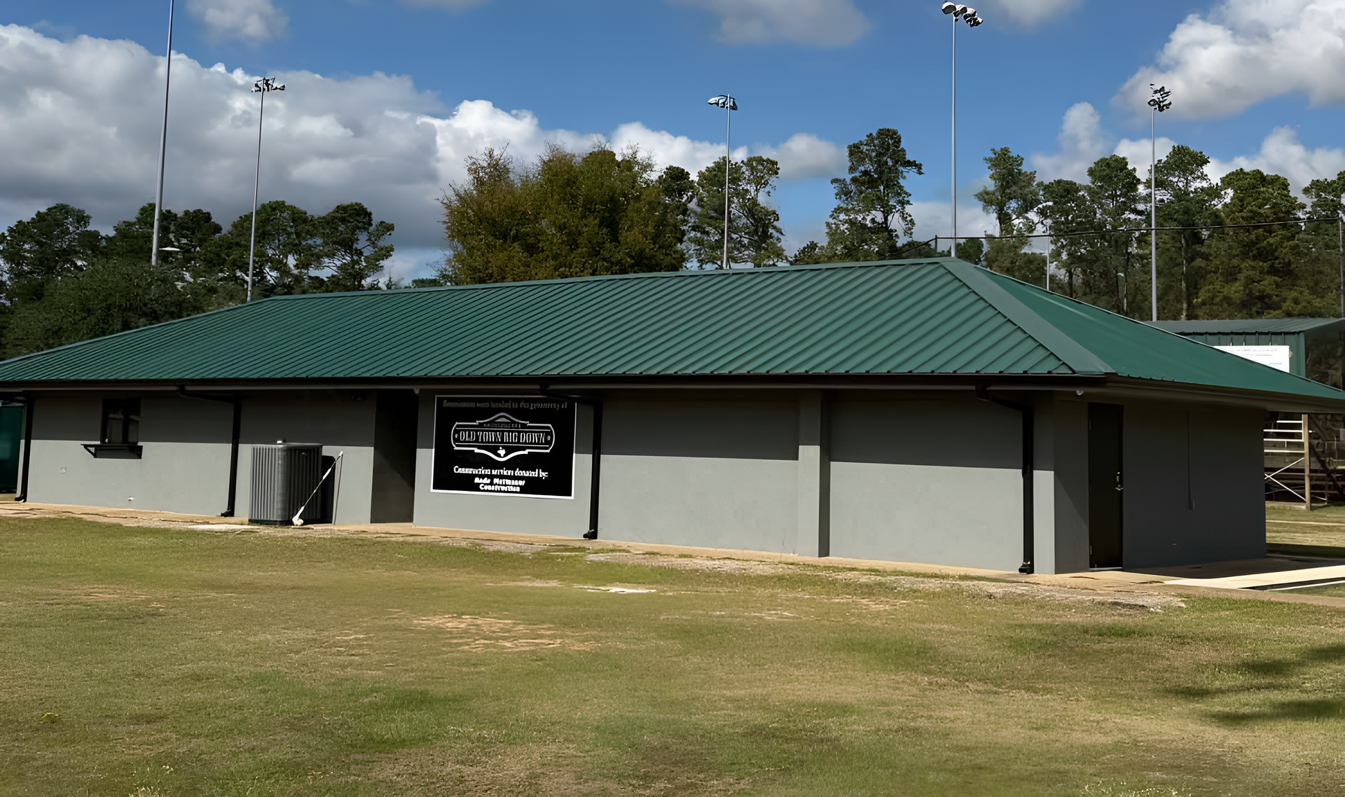 Single-story building with green metal roof, white walls, and a sign, set on a grassy field under a partly cloudy sky