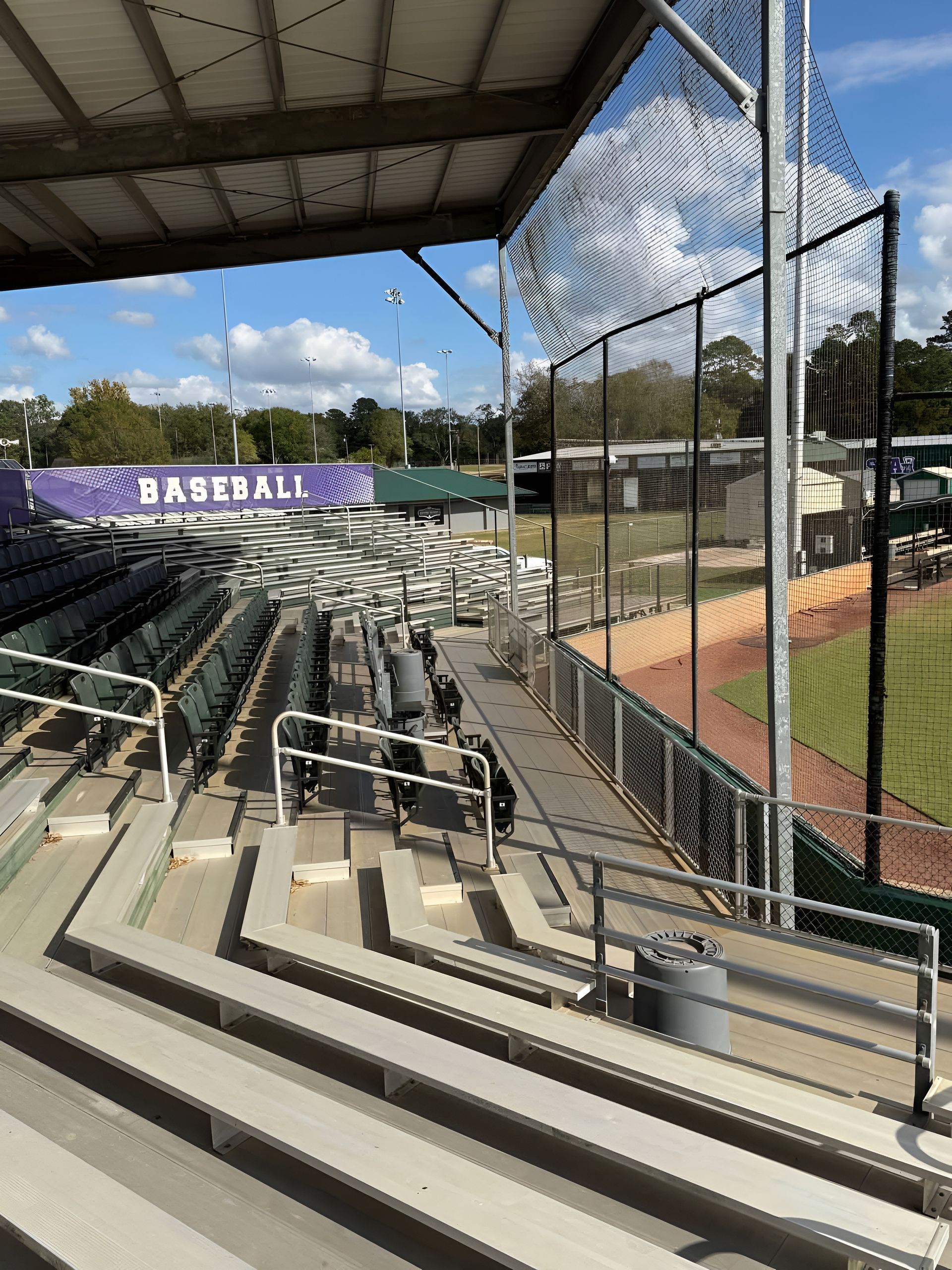 Empty baseball stadium bleachers under a roof, with a chain-link backstop and field beyond