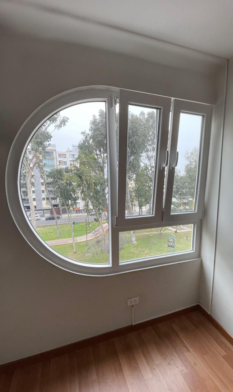 Una ventana blanca única, con secciones semicirculares y rectangulares, con vista a un parque de la ciudad.
