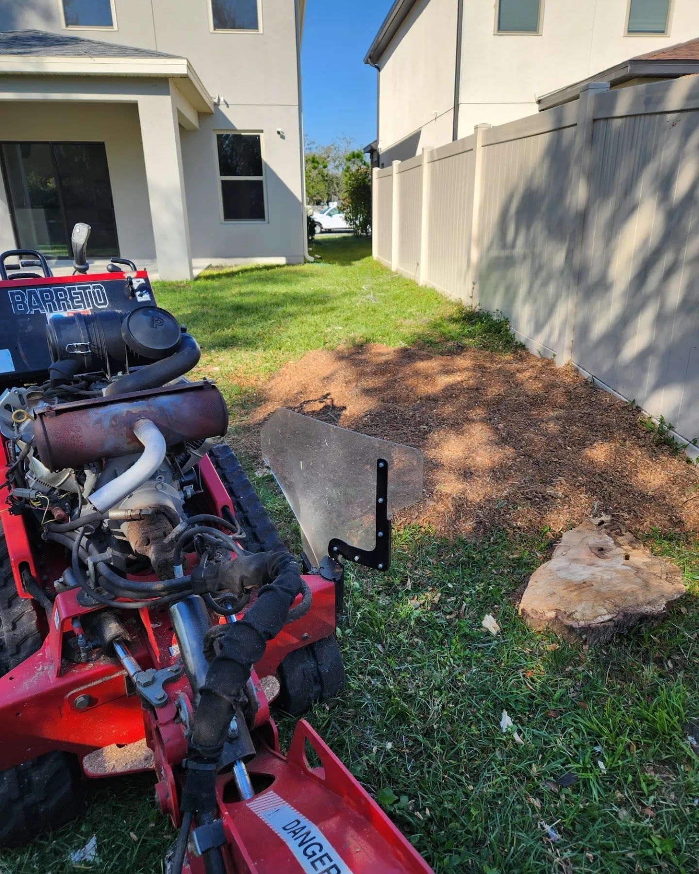 Red trencher machine next to a brown mulch area in a yard between two houses and a fence.