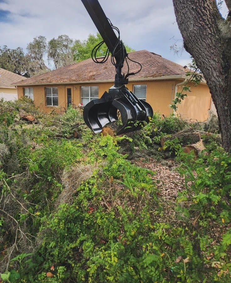 Hydraulic grapple arm lifting tree debris in front of a yellow house with brown roof.