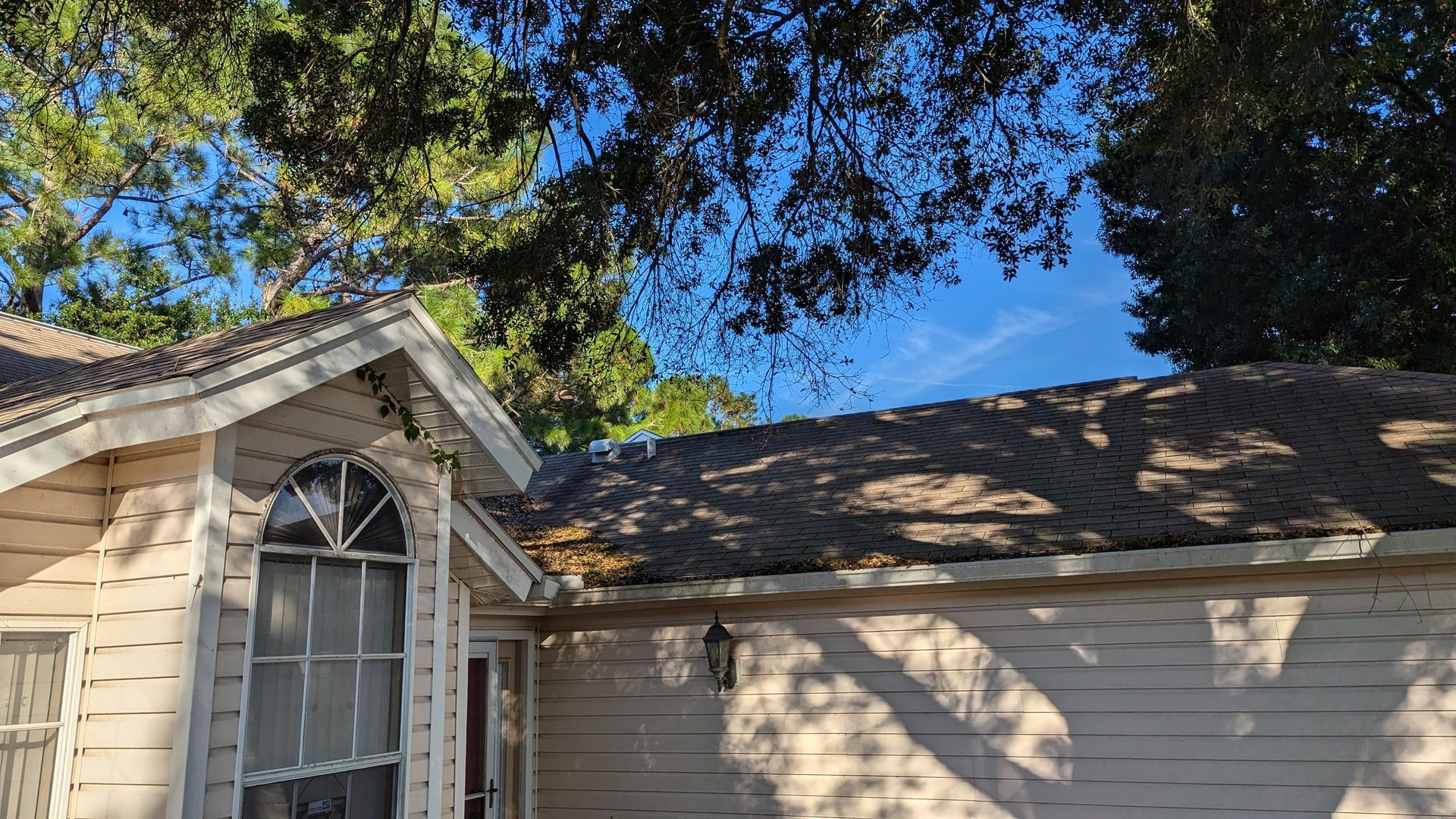 House with light-colored siding, brown roof, and arched window. Trees and blue sky visible.