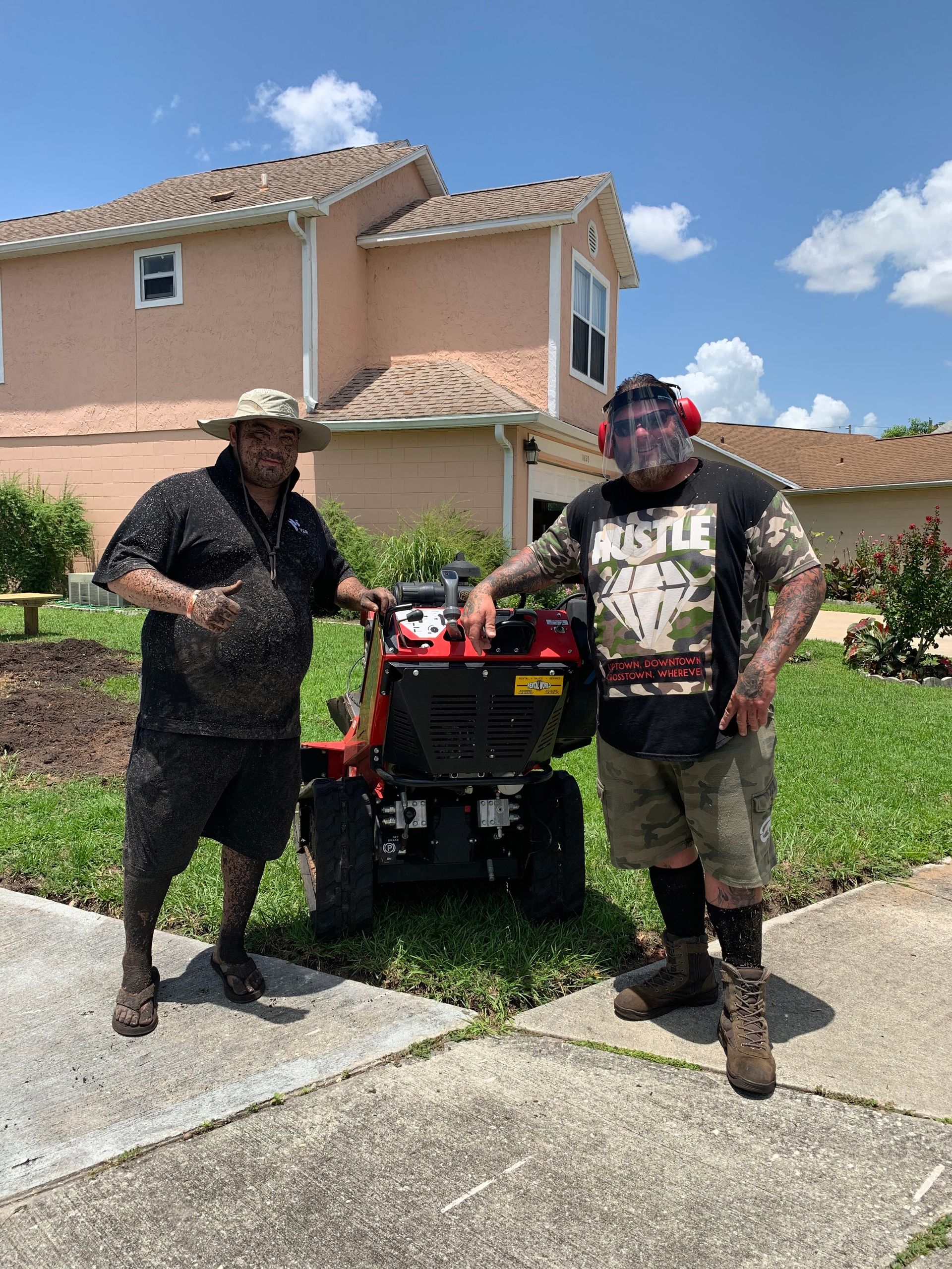 Two men pose with a red lawn aerator on a sunny day in front of a house.