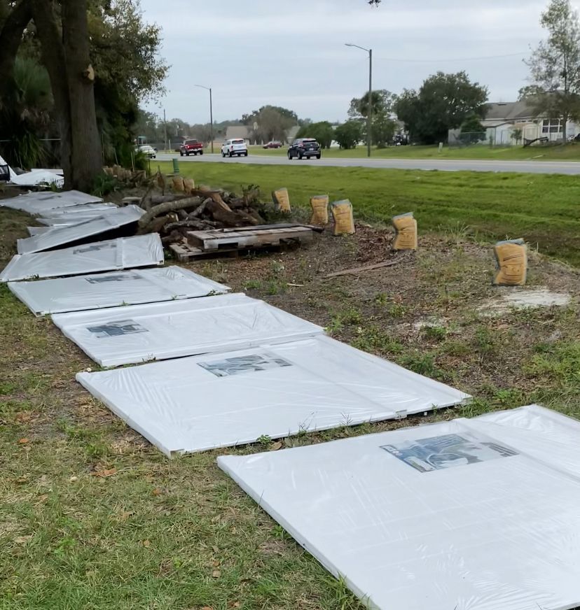 White panels laid on grass near road, next to tree and wood debris. Cars on road in the background.