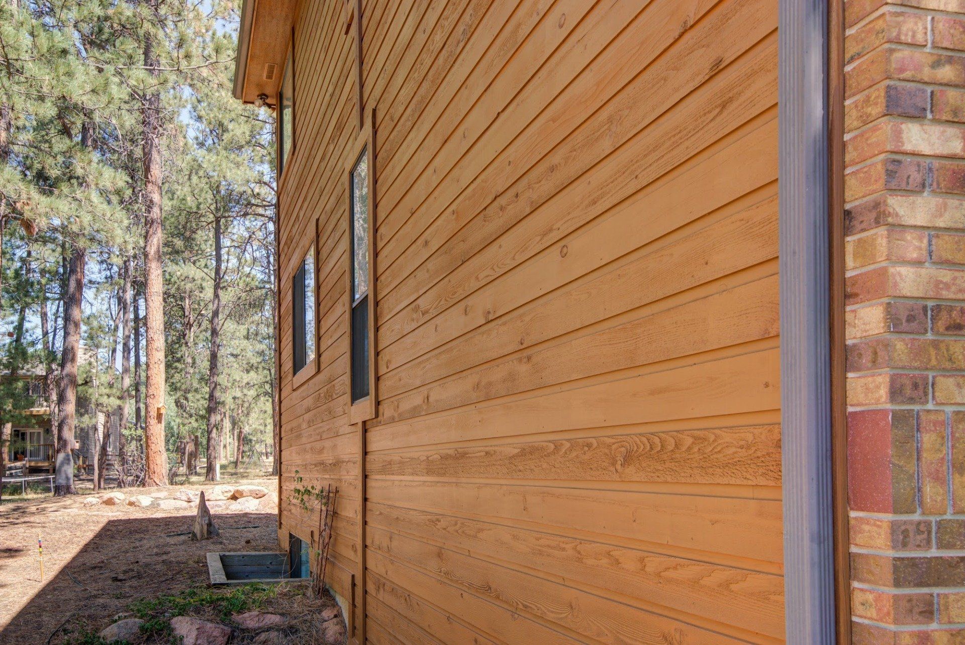 The side of a wooden house with a brick wall and trees in the background.